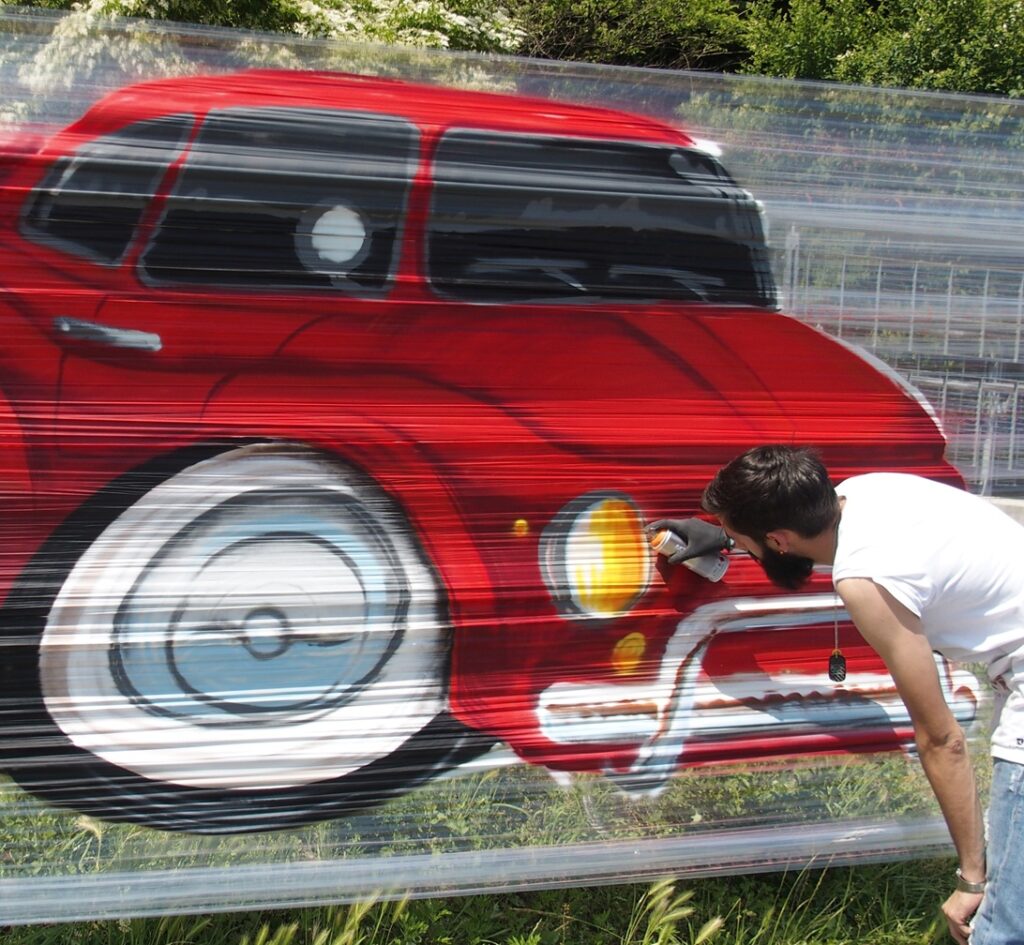 Primo piano delle mani di un writer che ritocca con la bomboletta spray il faro di un'auto dipinta su un murale